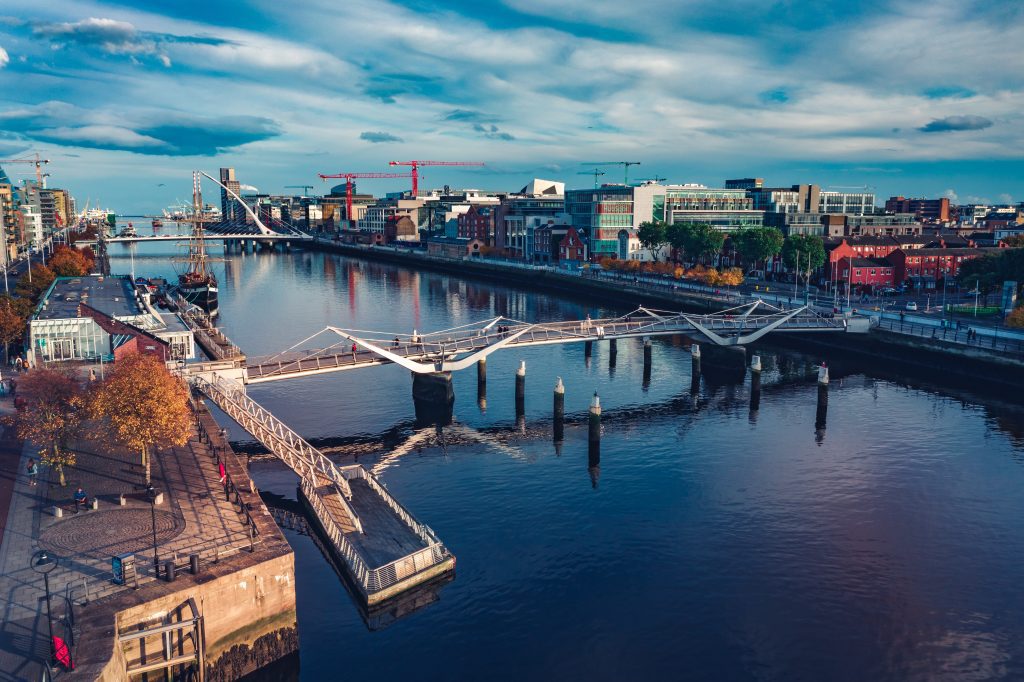 A modern pedestrian bridge spans a calm river in a vibrant urban setting under a dynamic sky.