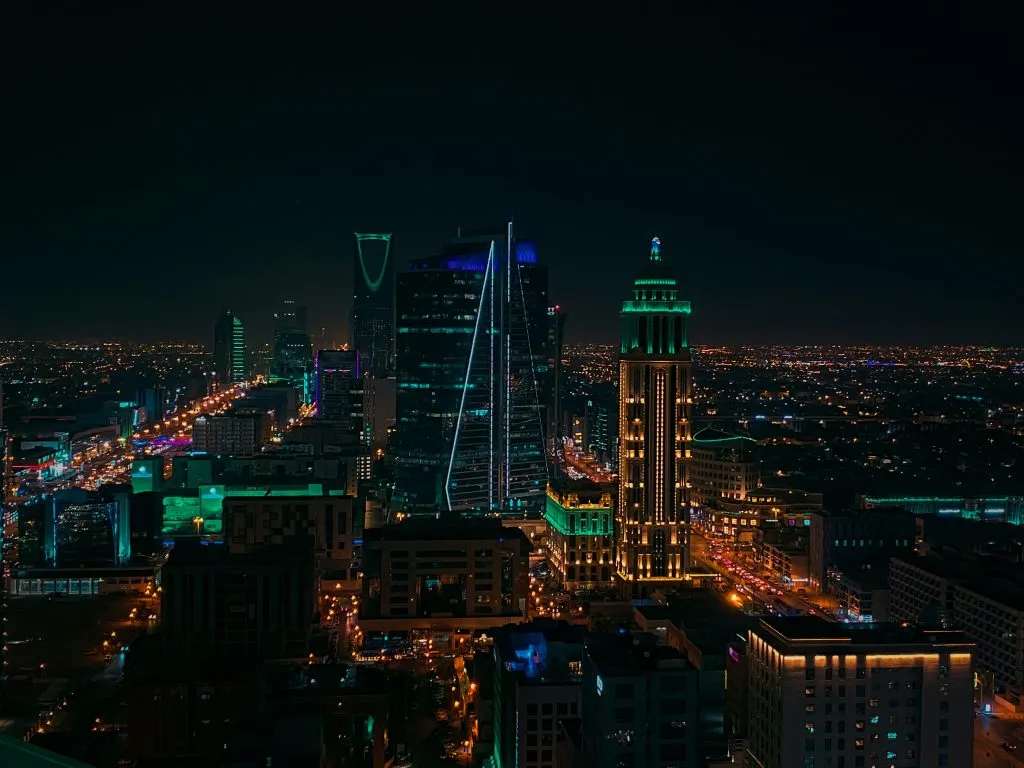 A vibrant nighttime cityscape of Riyadh, featuring illuminated skyscrapers and bustling streets.