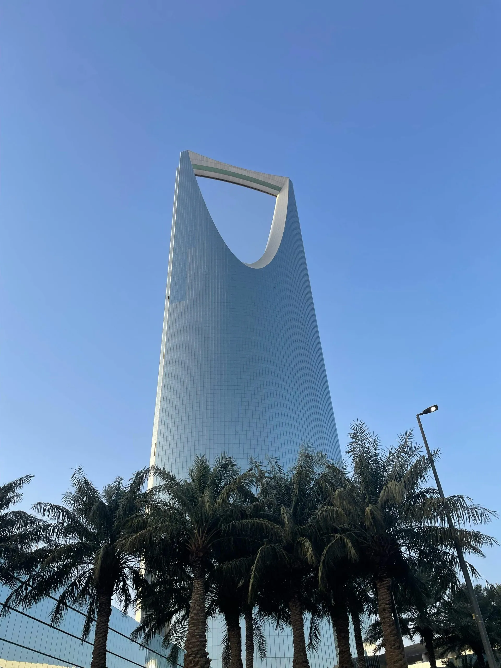 Kingdom Centre Tower in Riyadh, Saudi Arabia, framed by palm trees under a clear blue sky.