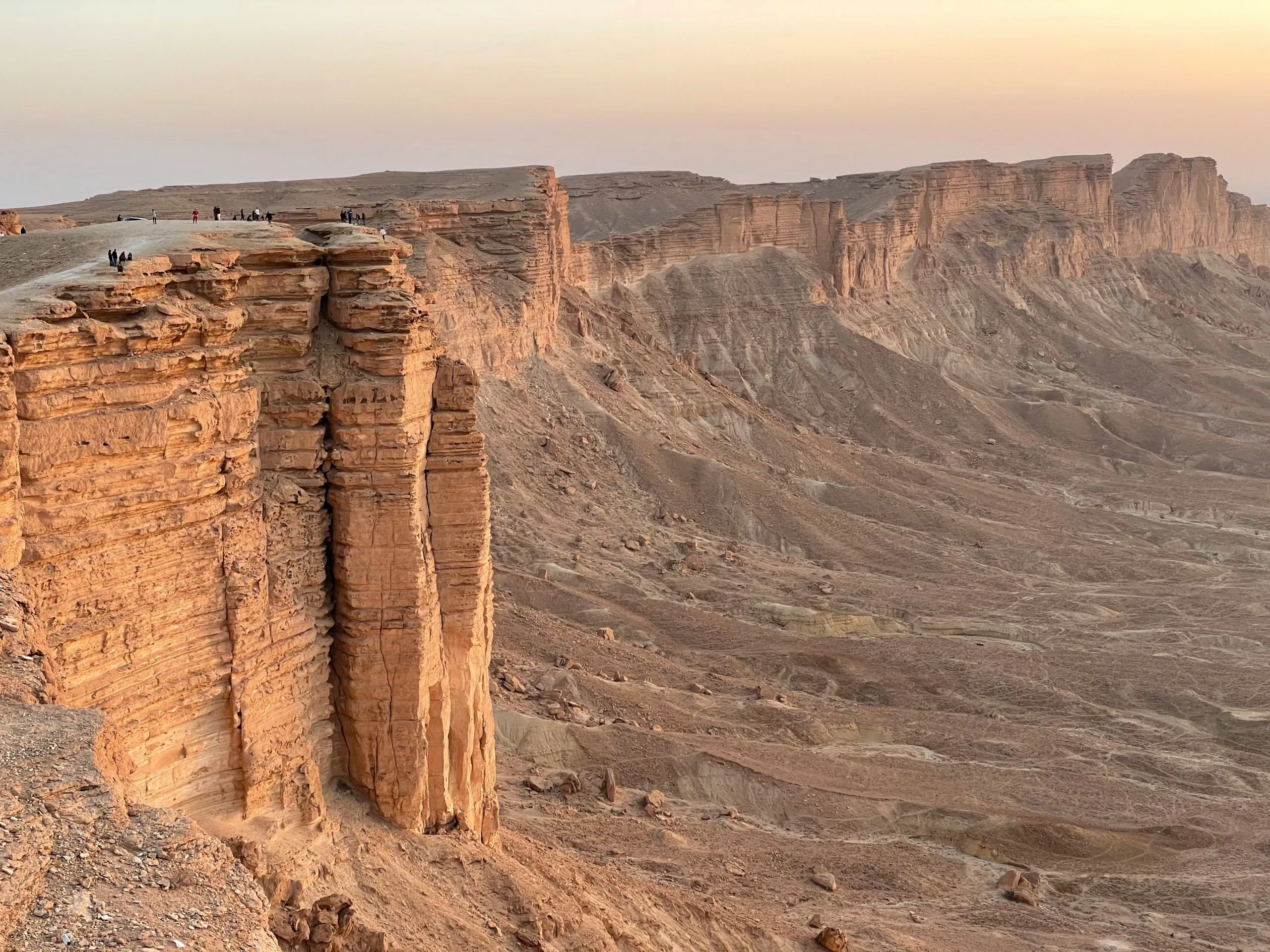 A dramatic desert cliff formation with visitors standing near the edge, illuminated by soft sunlight.