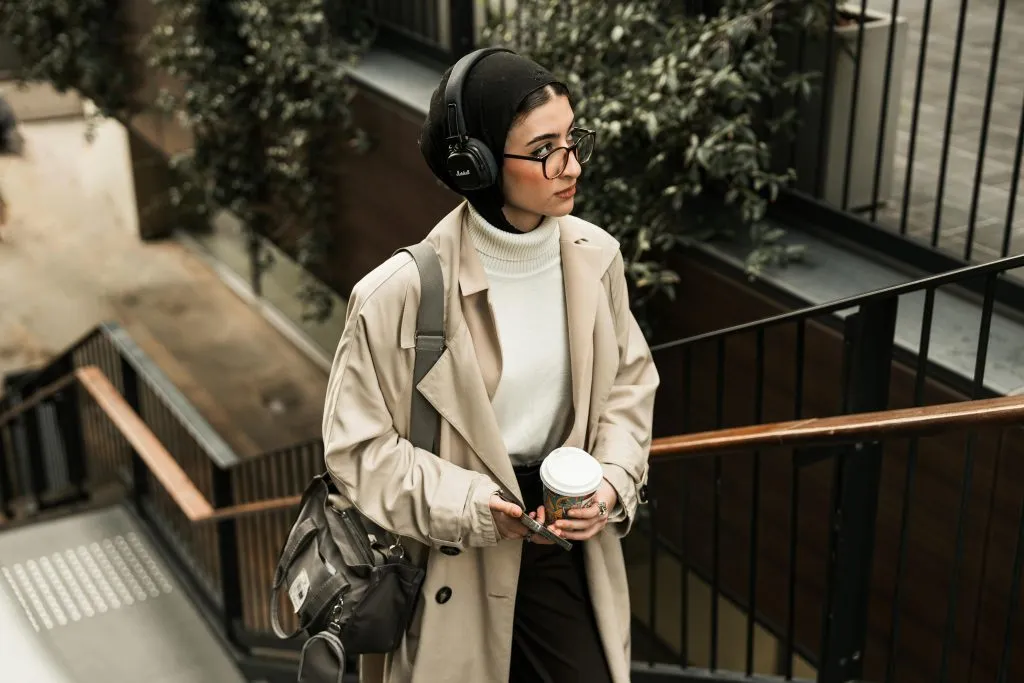A person in a beige trench coat and headphones holds a coffee cup while walking up stairs outdoors.