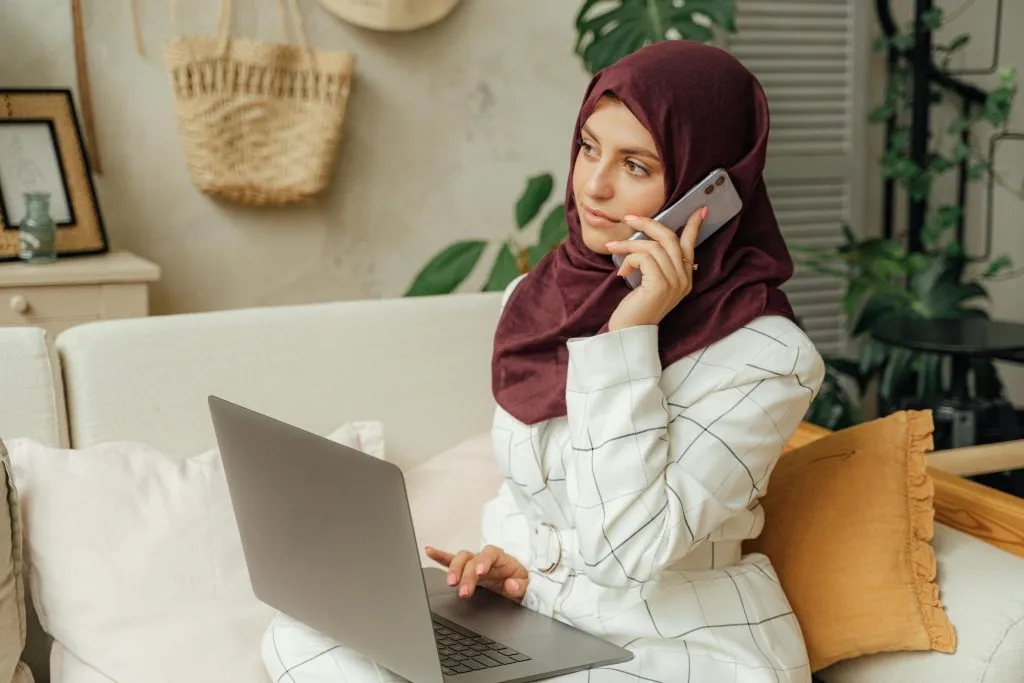 A woman in a burgundy hijab works on a laptop while speaking on the phone in a cozy setting.