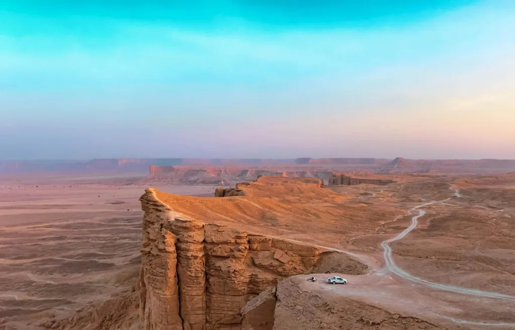 A vast desert landscape with cliffs, a winding road, and a parked car under a gradient sky.