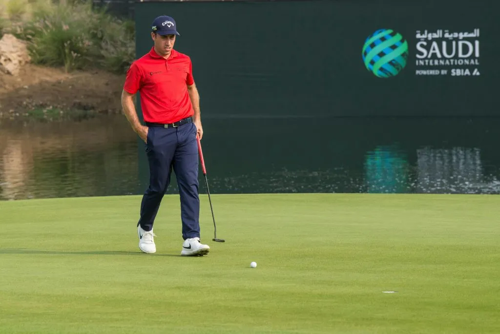 A golfer in a red shirt and navy pants prepares to putt on a green near a Saudi International sign.