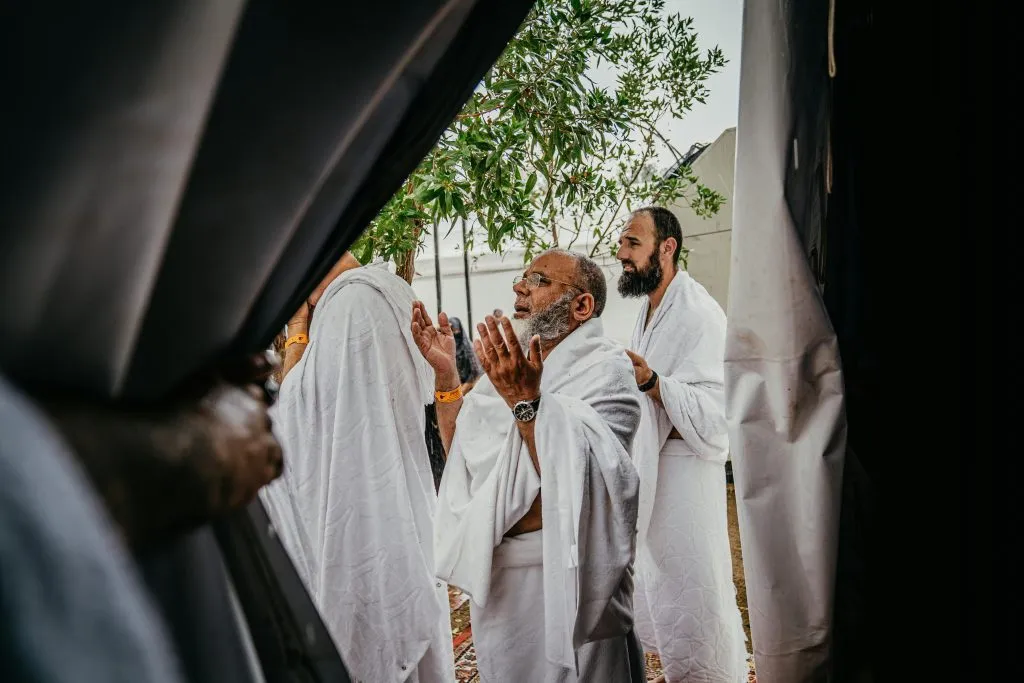 Men dressed in white Ihram garments stand outdoors, engaged in prayer during a religious ritual.