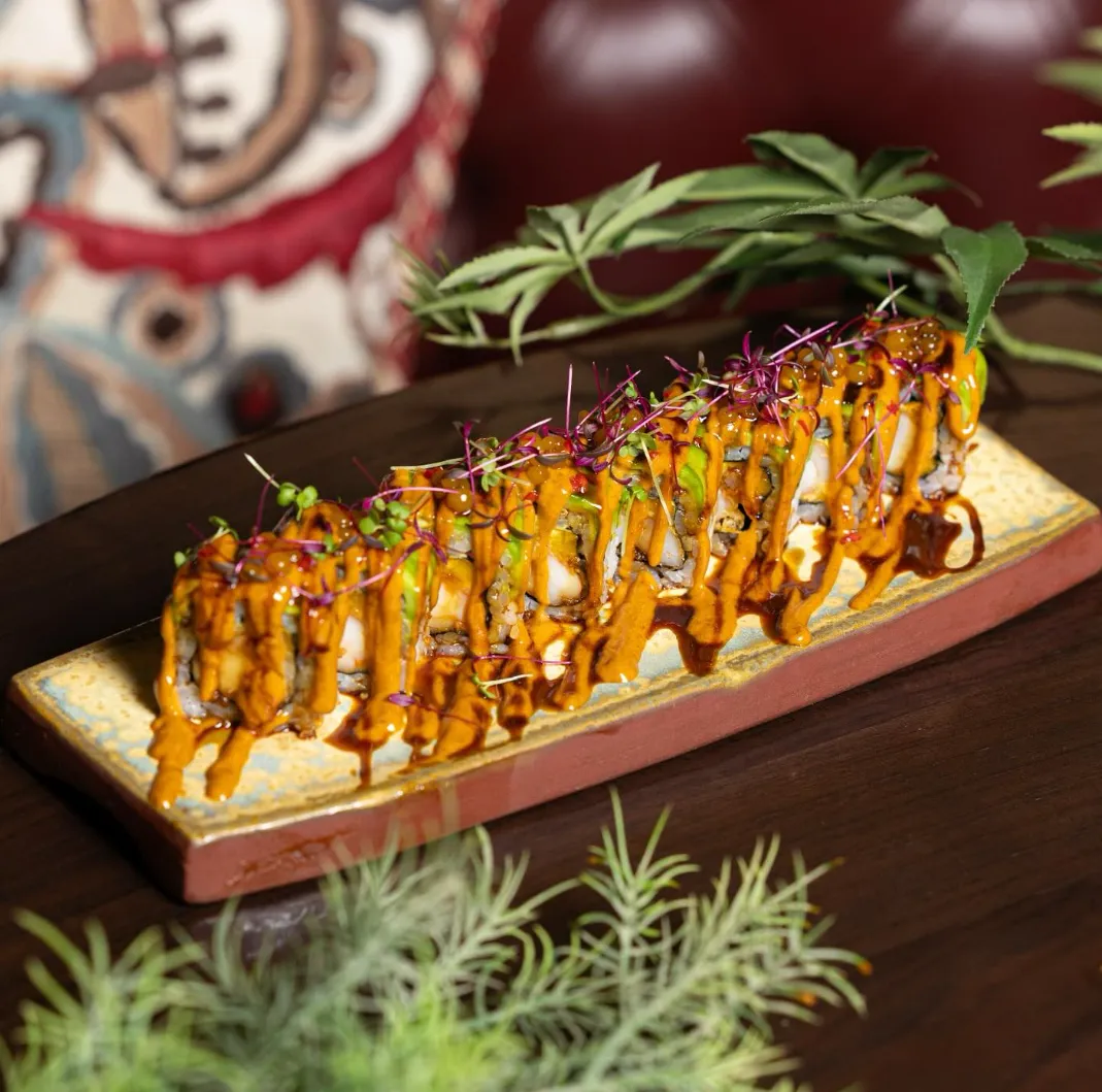 A plated sushi roll topped with vibrant sauces, microgreens, and garnishes on a wooden table.