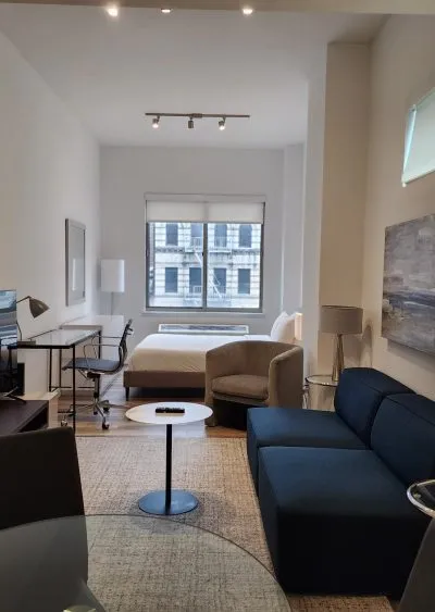 Modern studio apartment with a bed, desk, sofa, and large window, featuring minimalist decor.