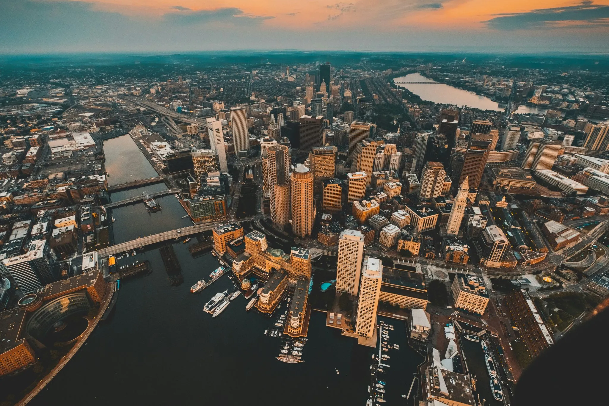 Aerial view of Boston's skyline at sunset, featuring skyscrapers, waterways, and urban architecture.