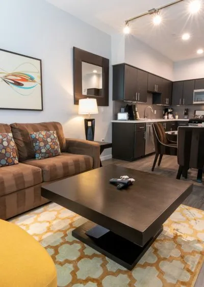 Modern living room with brown furniture, geometric rug, and open kitchen in the background.