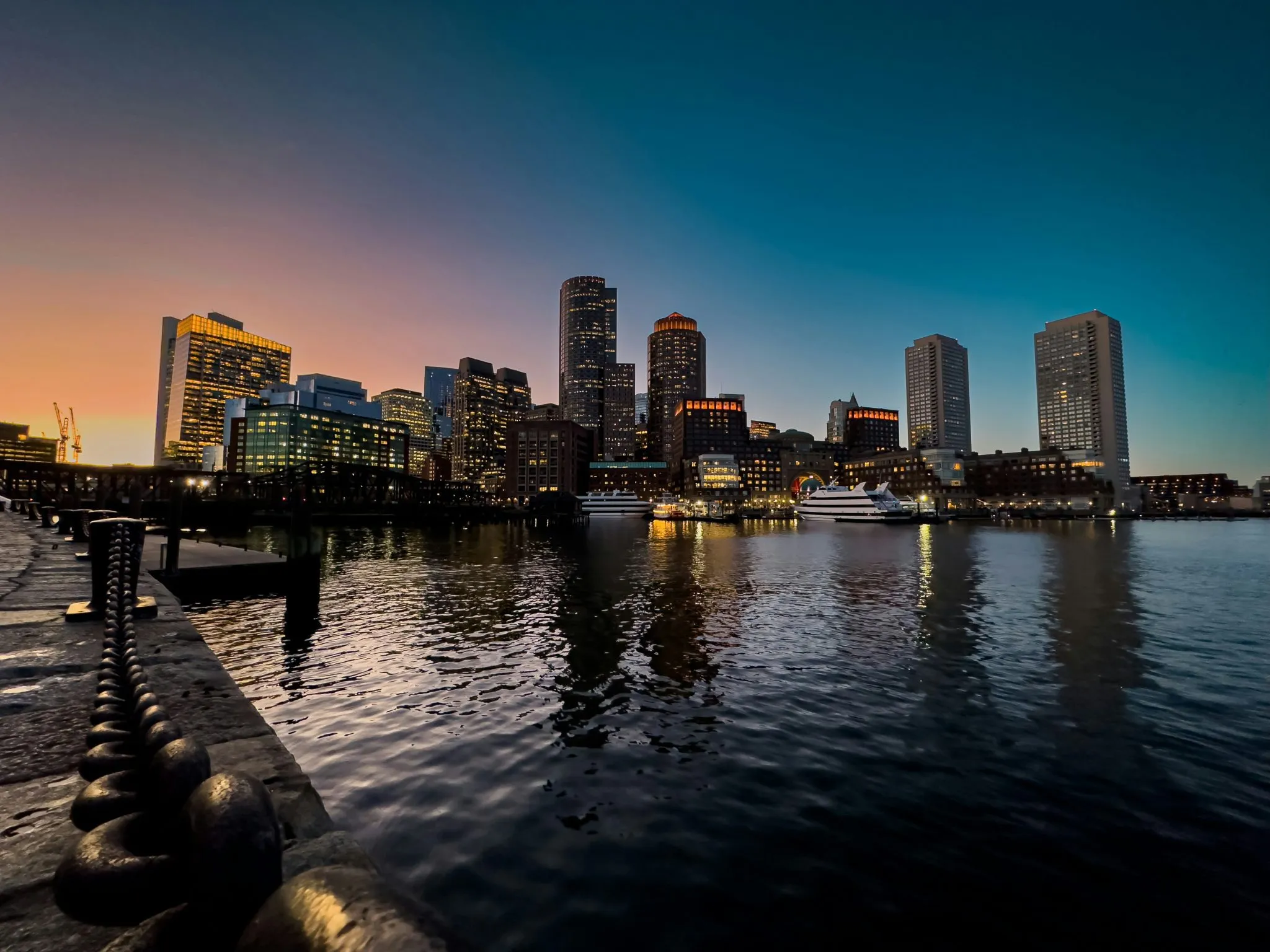 A waterfront cityscape at dusk, featuring illuminated skyscrapers and calm reflective waters.