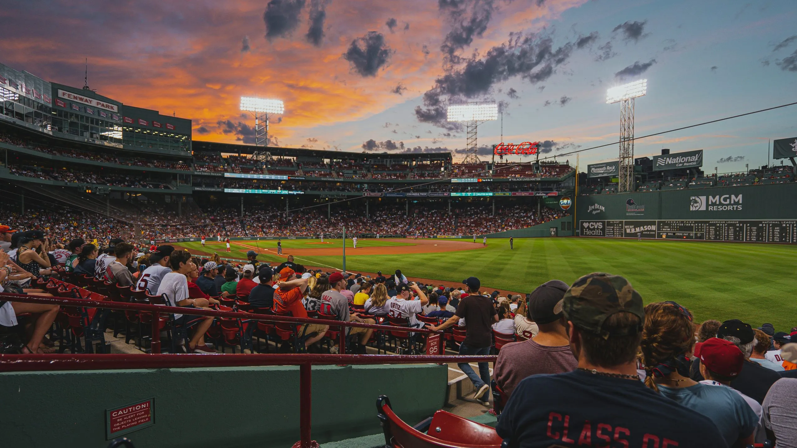 Fenway Park at sunset, with fans watching a baseball game and the iconic Green Monster in view.