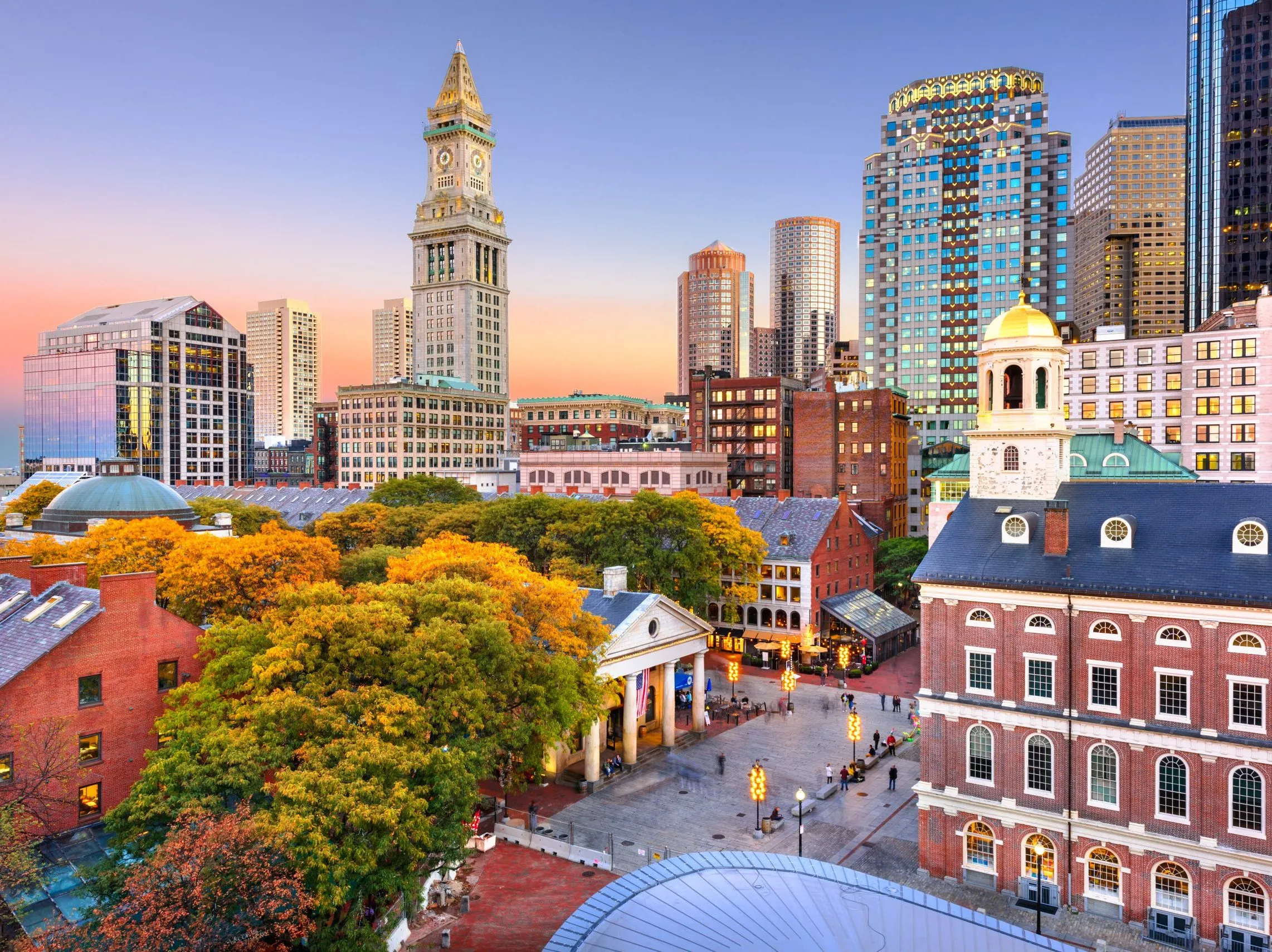 Boston skyline at sunset with historic Faneuil Hall and Quincy Market in the foreground.