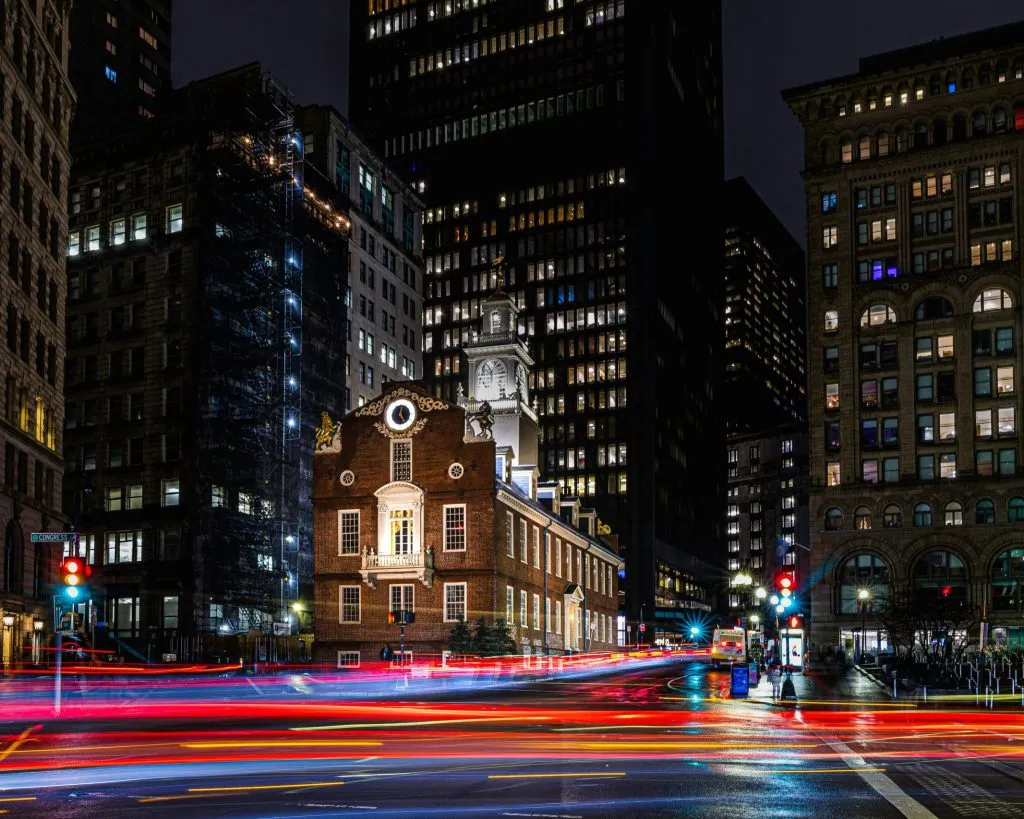 Historic Old State House illuminated at night, surrounded by modern skyscrapers and light trails.