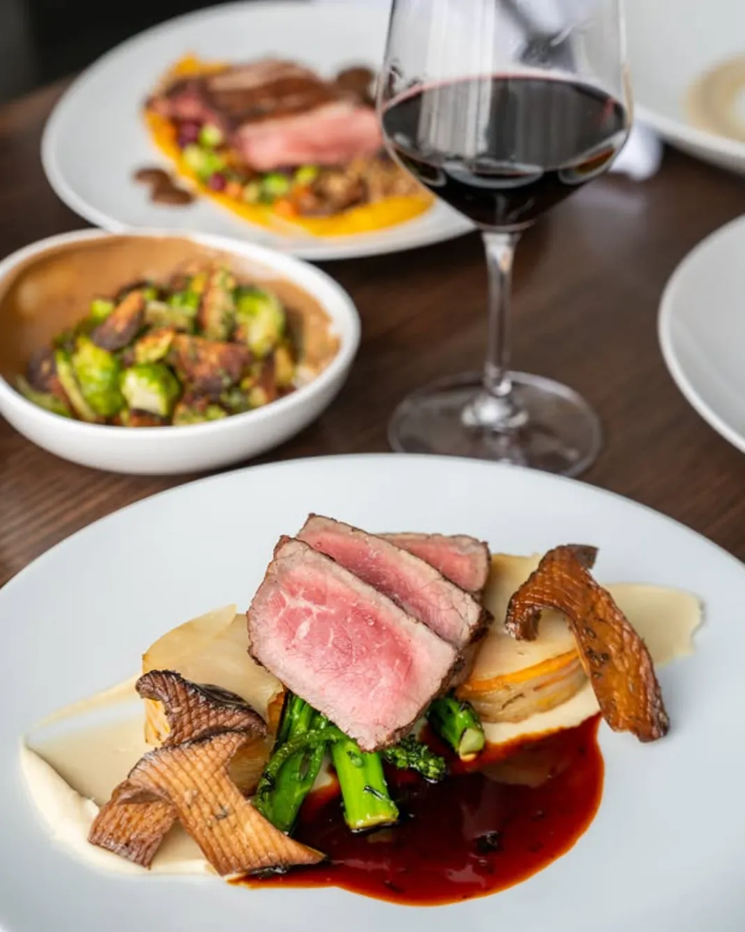 A gourmet dish featuring seared meat, vegetables, mushrooms, and red wine sauce, paired with wine.