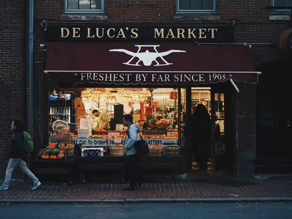 De Luca's Market storefront displays fresh produce and goods under a maroon awning on a brick street.