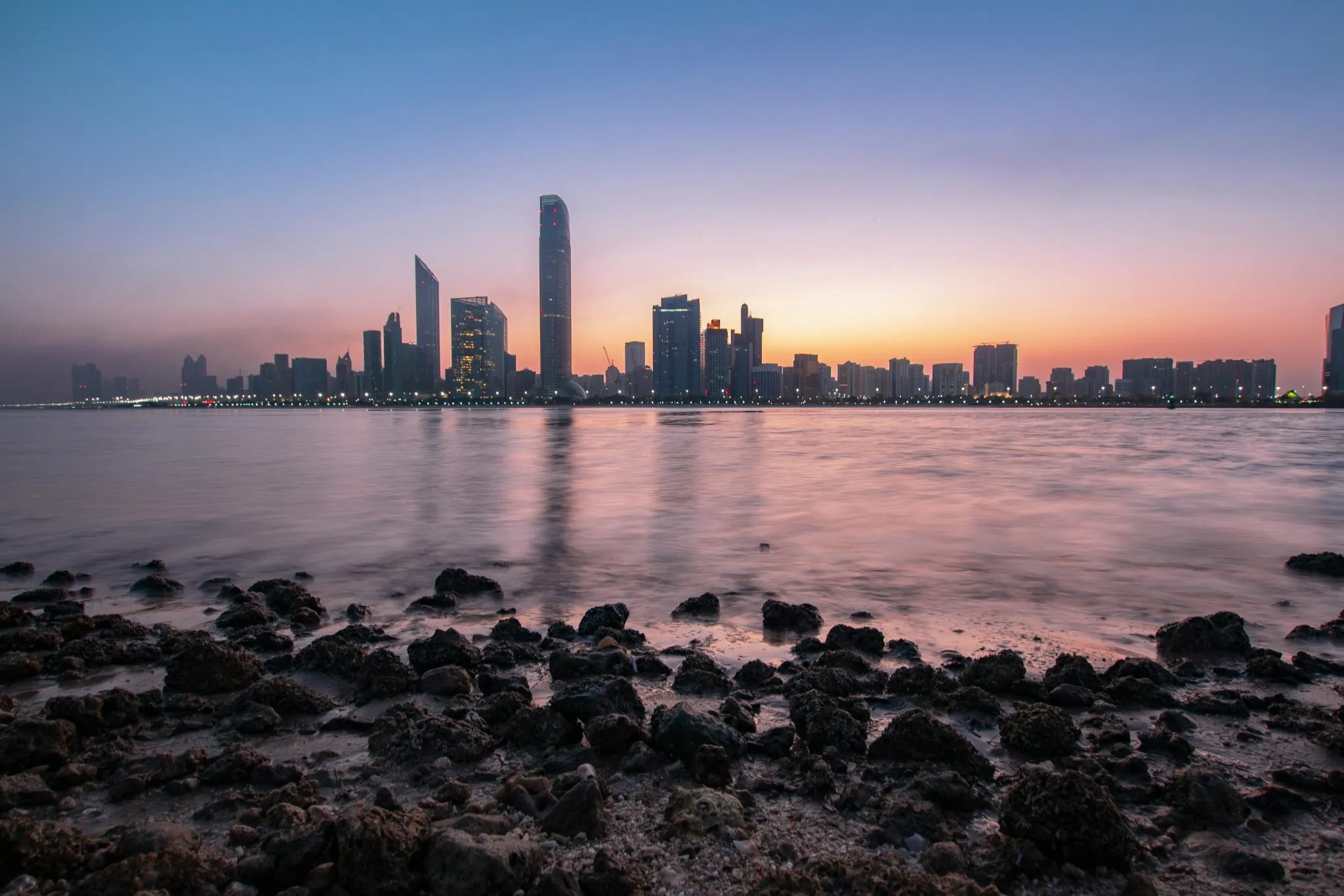 Abu Dhabi skyline at sunset, with calm waters and rocky foreground creating a serene urban scene.