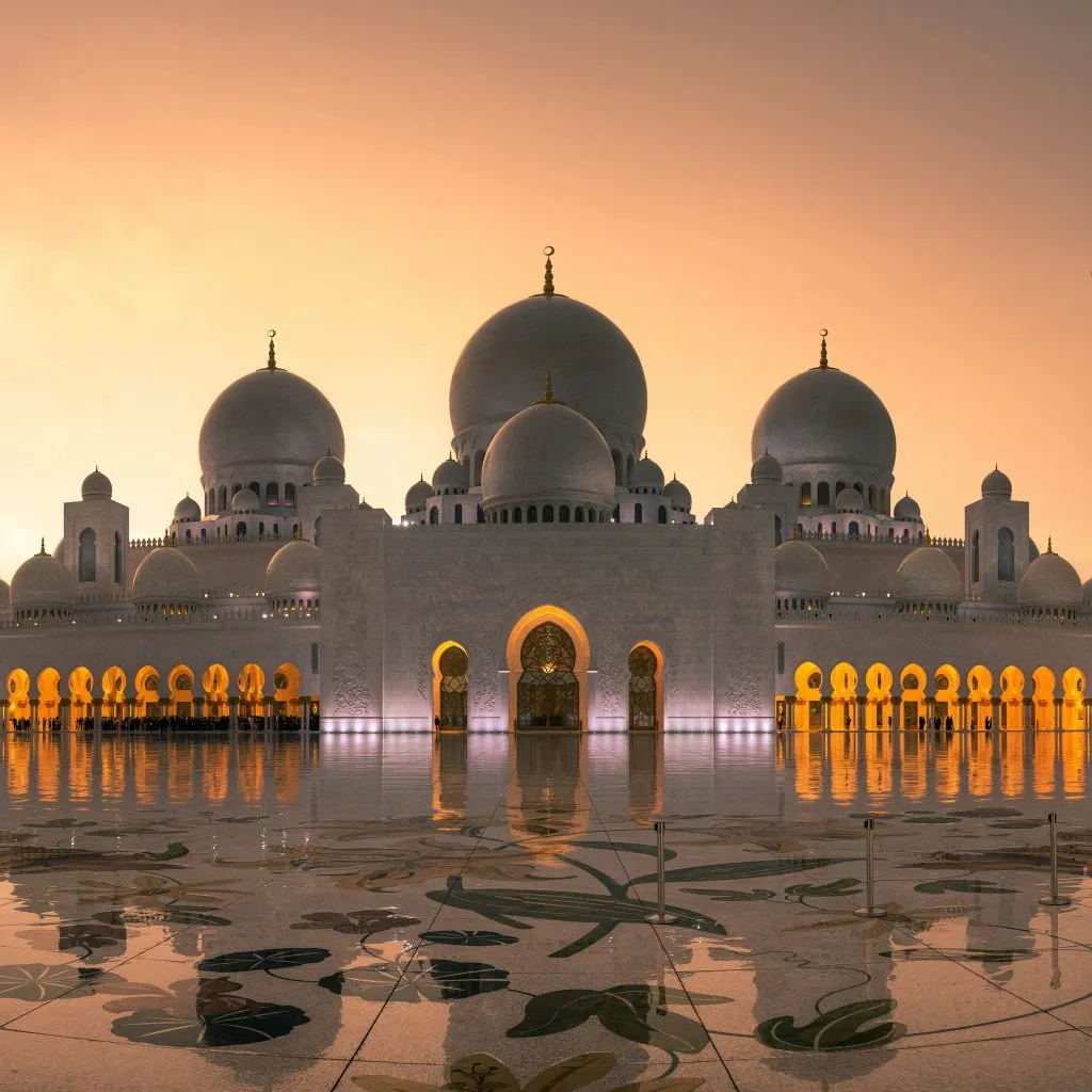 Grand mosque with domes and arches, reflecting warm sunset hues on polished marble flooring.
