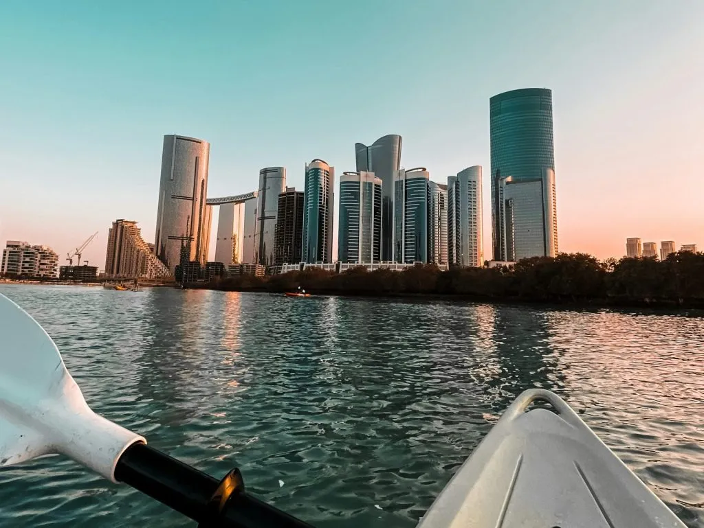 A modern cityscape with skyscrapers reflecting sunlight, viewed from a kayak on calm waters.