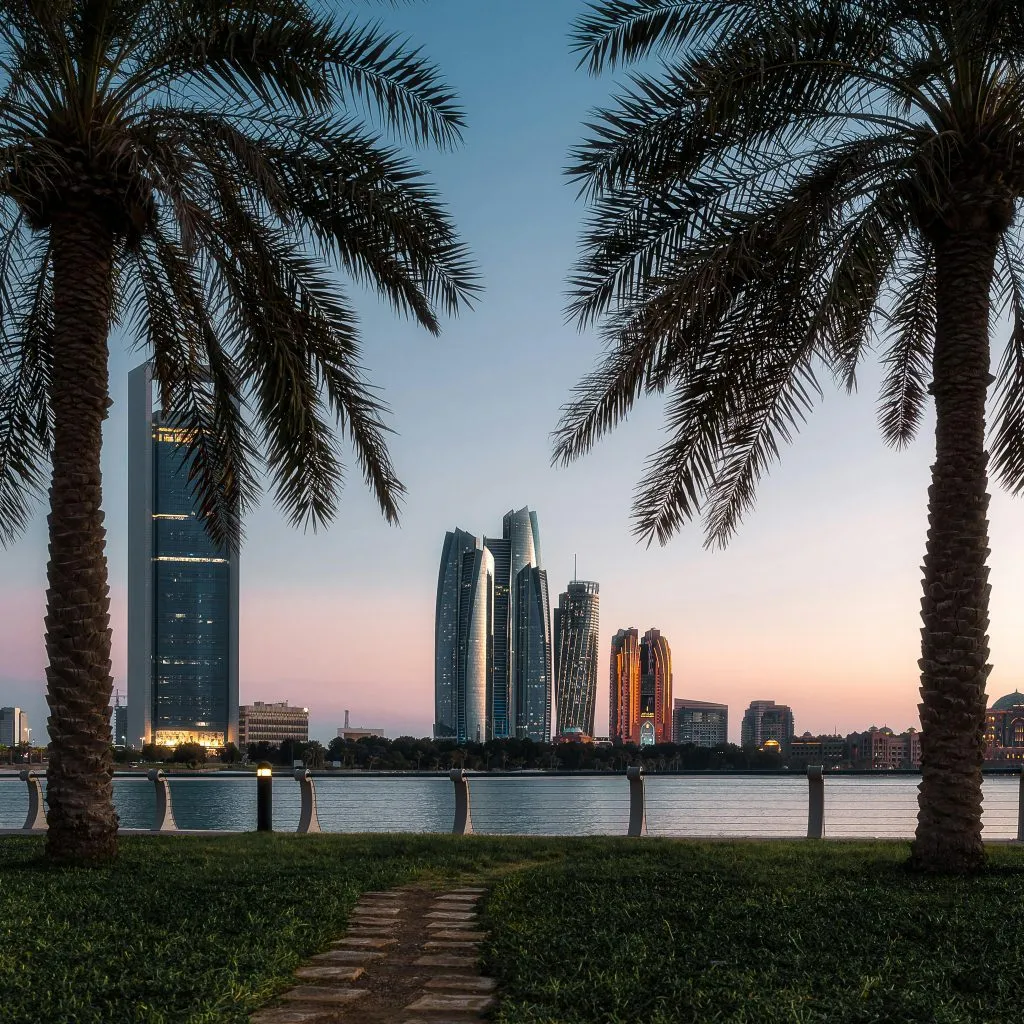 Palm trees frame a waterfront view of modern skyscrapers at sunset in Abu Dhabi, UAE.