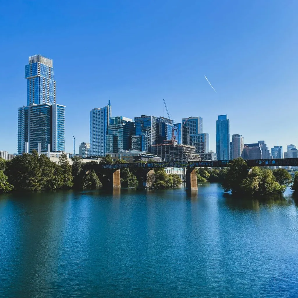 A modern city skyline with tall buildings, a bridge, and a calm river under a clear blue sky.