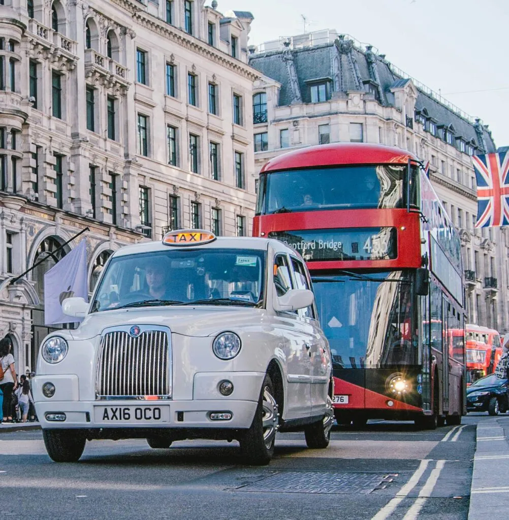 A white London taxi and a red double-decker bus navigate a busy street with historic architecture.