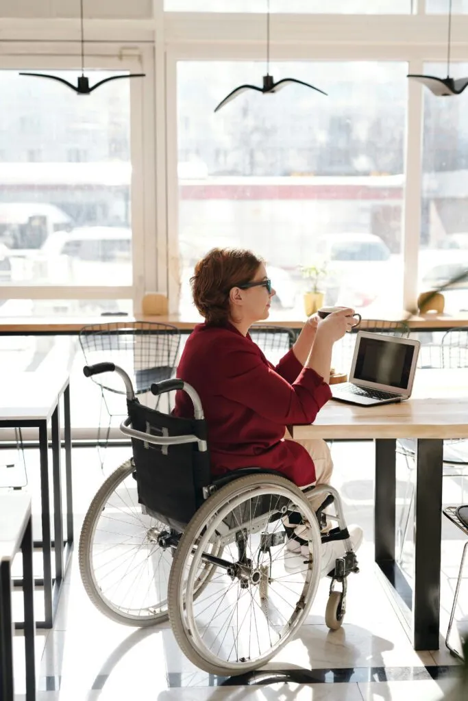 A woman in a wheelchair holds a cup while seated at a table with a laptop in a bright café.