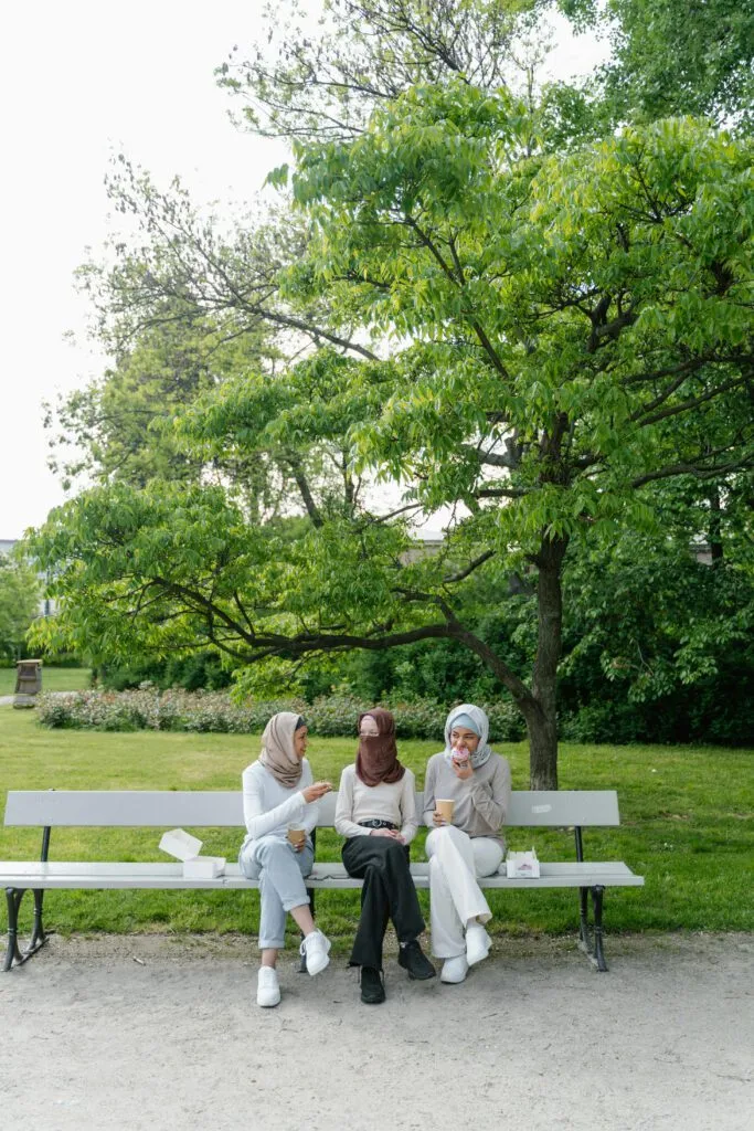 Three women in hijabs sit on a park bench, conversing and holding coffee cups amidst greenery.