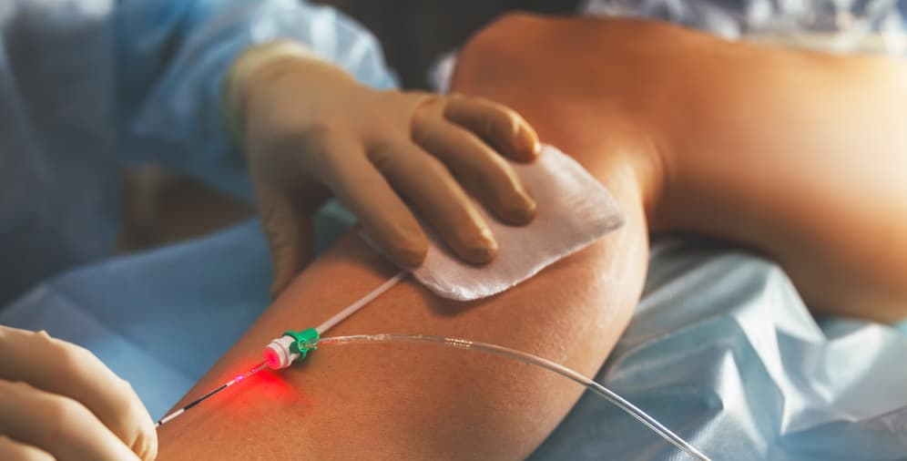 A gloved hand placing a small medical adhesive bandage on a patient's arm, which has a catheter inserted and a red light indicating an active connection.