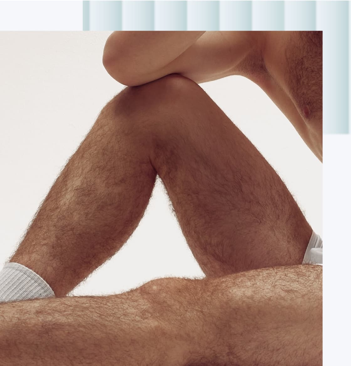 A close-up of two male legs, one crossed over the other, showing hair and skin texture. The legs are partially covered with light-colored underwear.