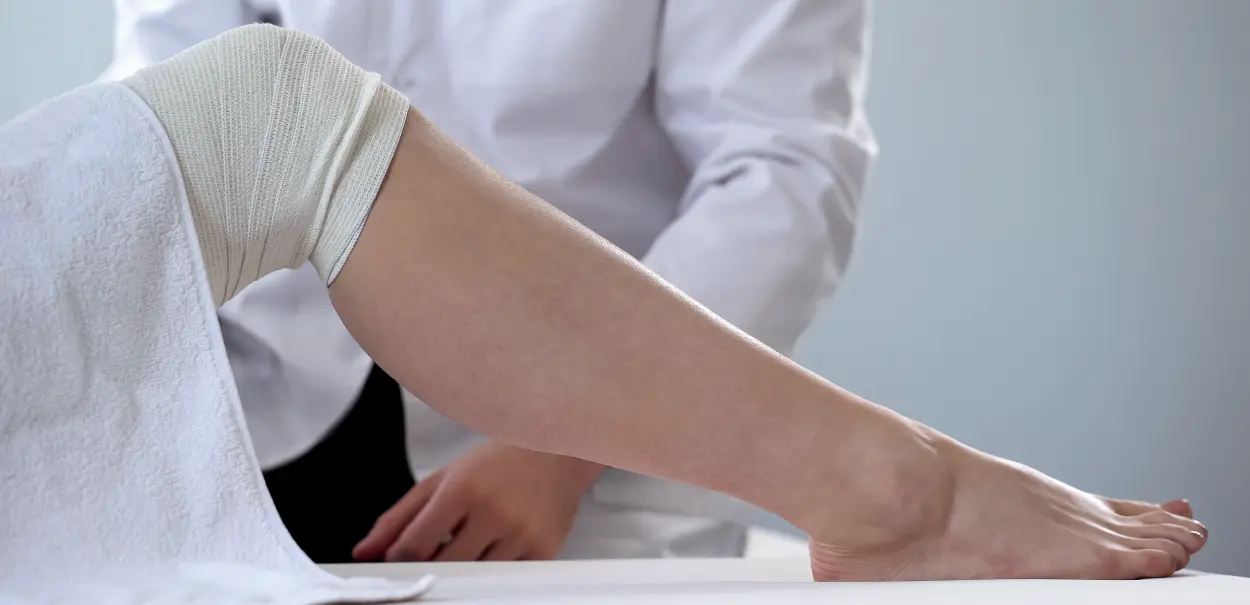 A person's leg wrapped in a bandage, lying on a table, with a hand of a medical professional adjusting the bandage.