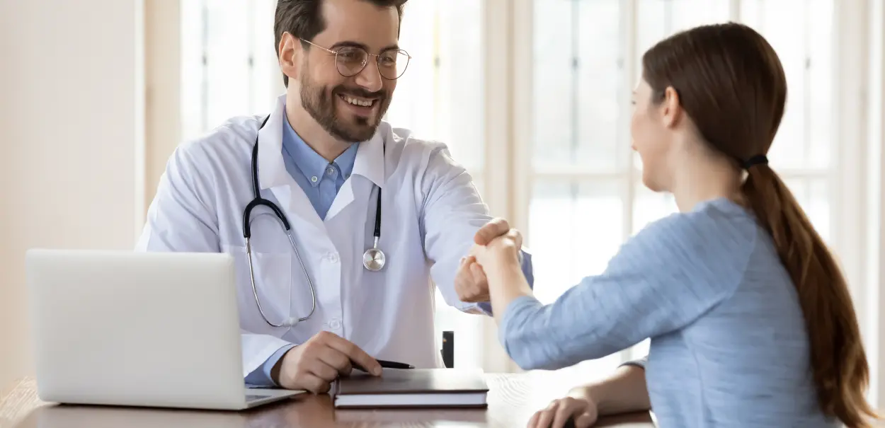 A smiling male doctor in a white coat, holding a pen and a clipboard, shakes hands with a female patient who is seated across from him at a desk with a laptop on it.