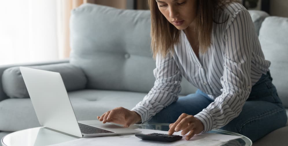 A woman in a striped shirt using a calculator while working on a laptop, with documents spread out on a glass table.