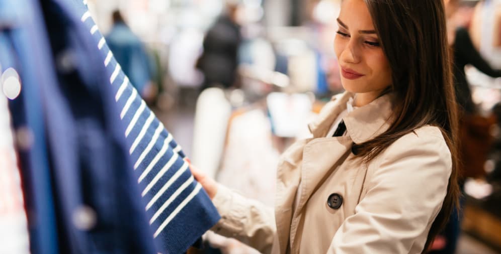 A woman with long dark hair is holding up a blue sweater with white stripes while she is shopping in a clothing store.