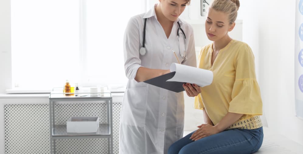 A female doctor wearing a white lab coat and stethoscope is reviewing a document with a female patient who is sitting on an examination table. The patient is wearing a yellow blouse and appears to be engaged in the conversation.