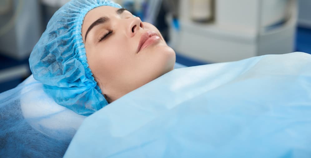 A woman lying down with her eyes closed, wearing a blue surgical cap and covered with a light blue surgical drape.