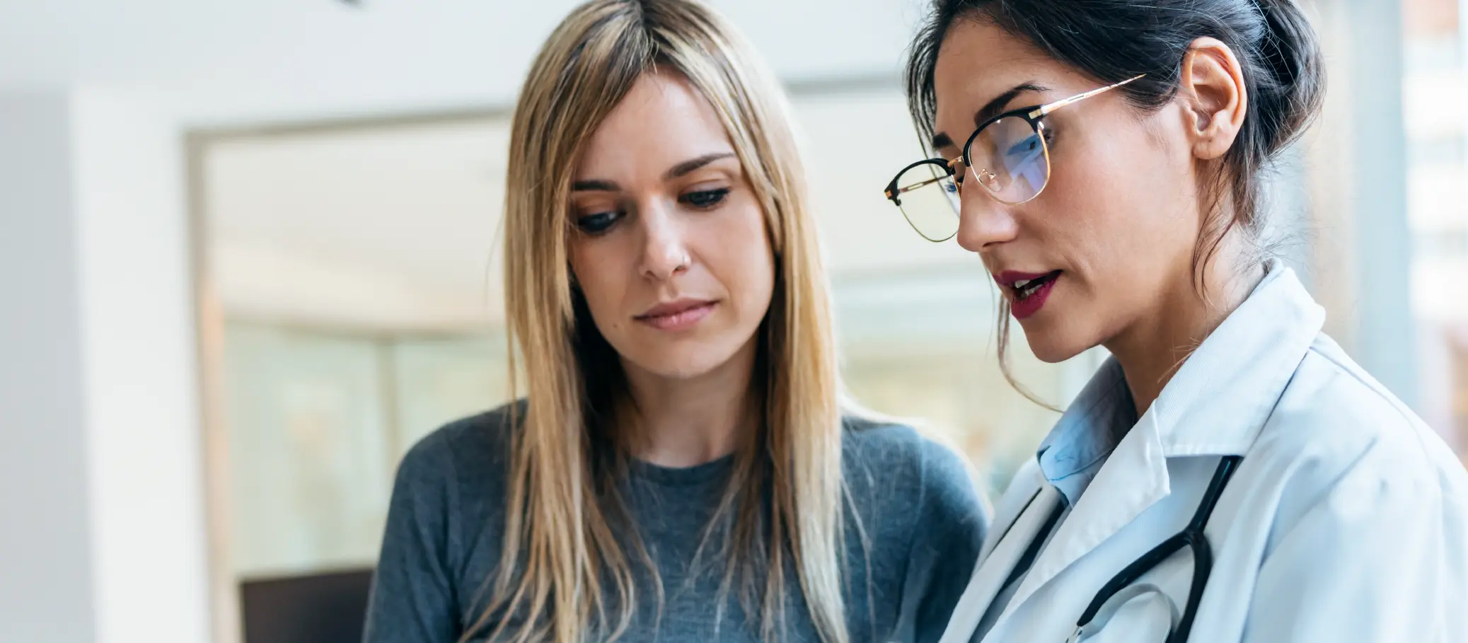 A woman in a doctor's coat with a stethoscope discussing something with another woman who is wearing a gray shirt.