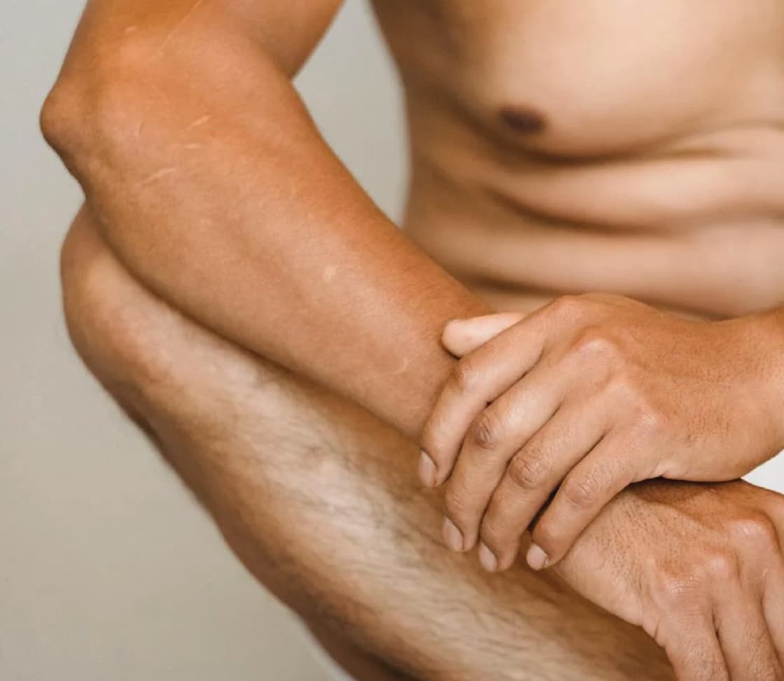 A close-up of a male torso with a focus on the arms and hands. The person is in a seated position with one hand resting on the knee and the other hand holding the wrist, showcasing a muscular build and natural skin tone.
