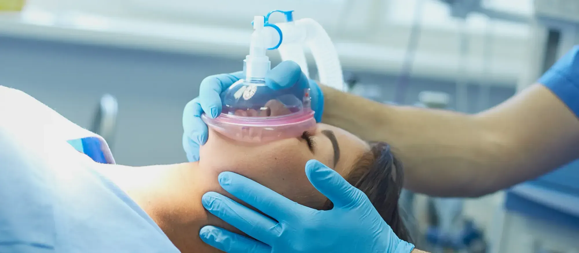 A medical professional wearing blue gloves holds a clear oxygen mask over a patient's face.