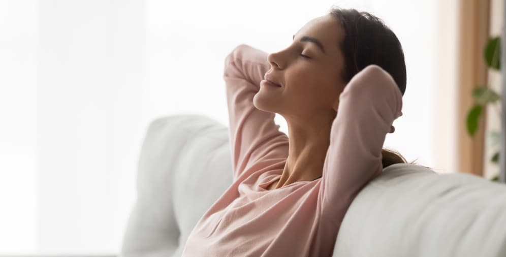 A woman sitting on a couch, relaxing with her eyes closed and hands behind her head, wearing a light pink long-sleeve top.