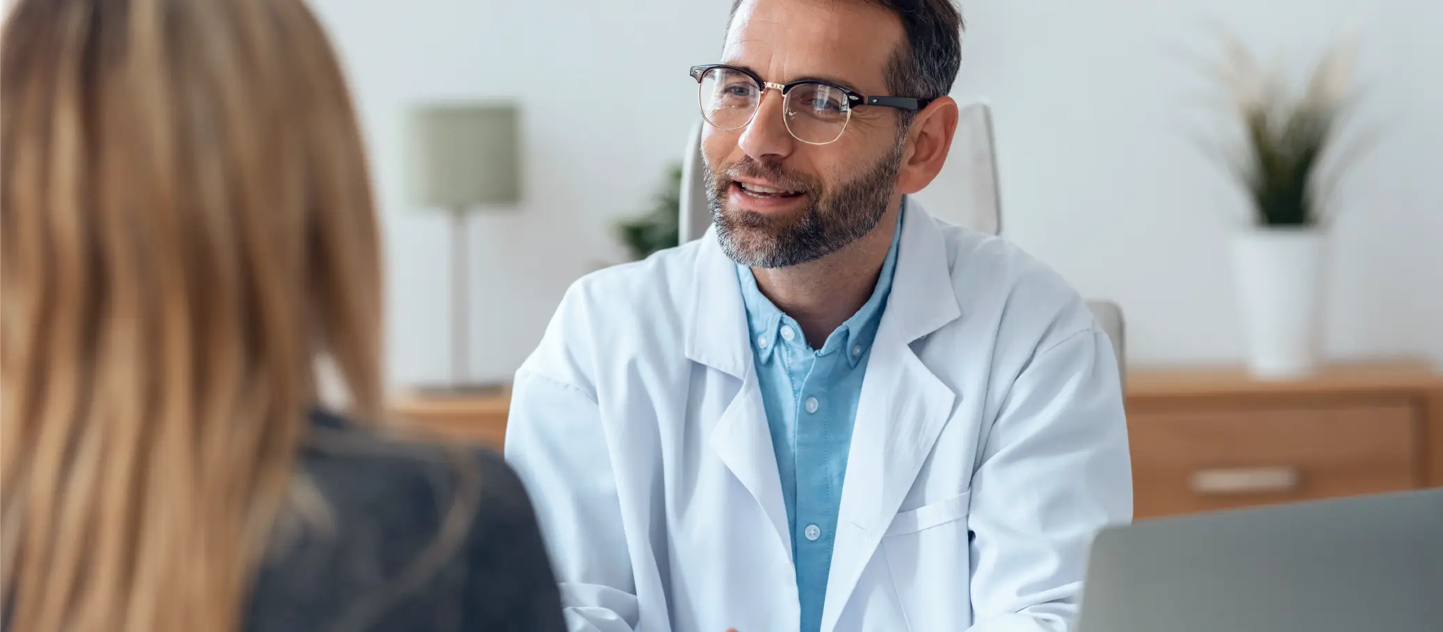 A male doctor in a white coat wearing glasses, smiling while talking to a patient. The doctor has a beard and short hair, and is seated at a table with a laptop and a plant in the background.