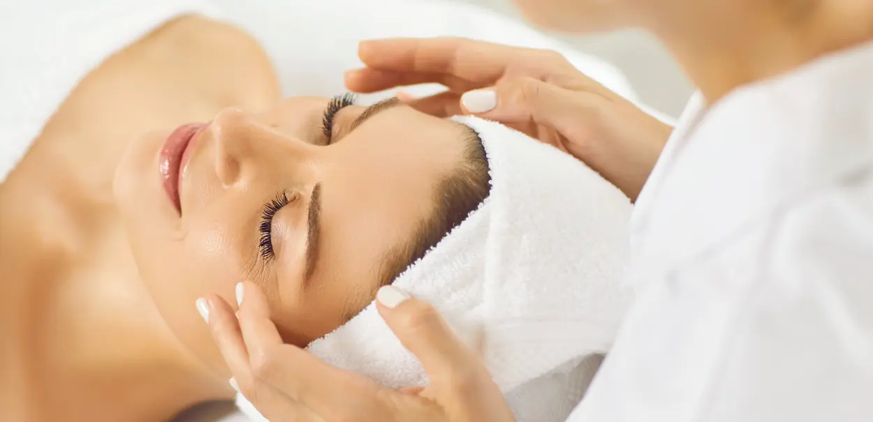 A woman receiving a facial treatment, relaxed with her eyes closed, as a person gently massages her head and temples.