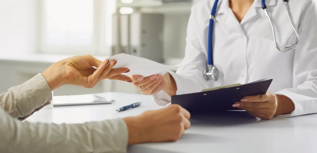 A healthcare professional in a white coat handing a document to a patient, who is holding a folder. The doctor has a stethoscope around their neck.
