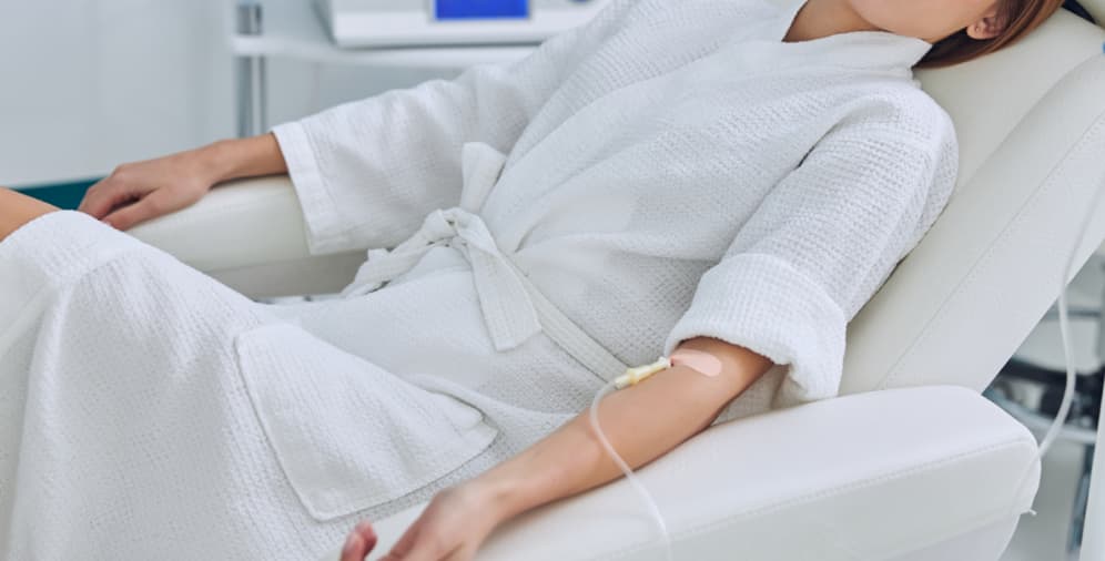 A person wearing a white robe reclines in a chair, with an intravenous drip attached to their arm.