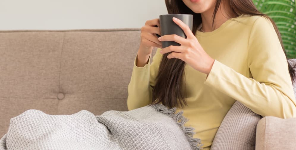 A woman wearing a yellow long-sleeve shirt holding a black mug, sitting on a couch with a gray blanket draped over her lap.