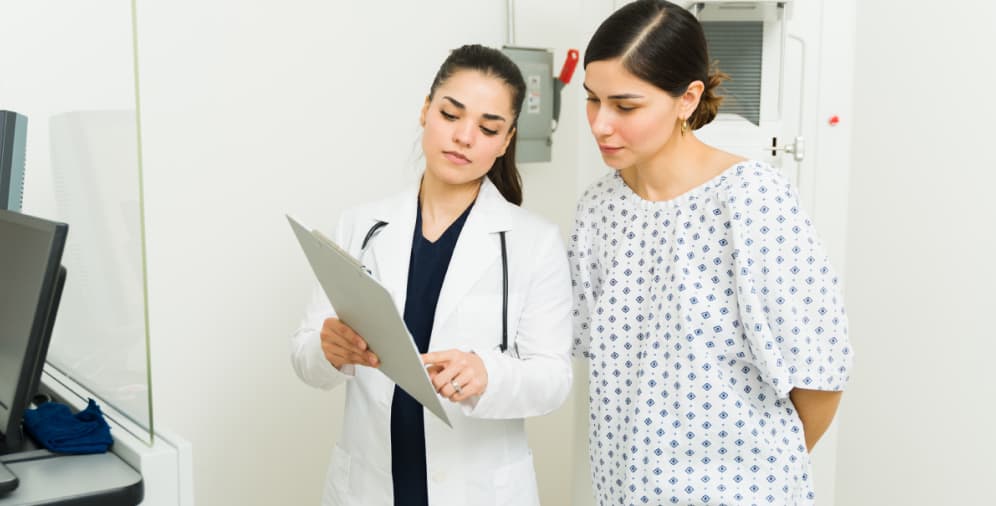 A healthcare professional in a white lab coat is reviewing a document with a patient, who is wearing a medical gown and showing a focused expression.