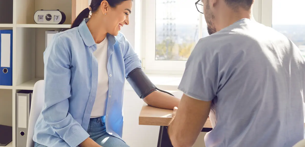 A woman sitting at a table smiling while having her blood pressure taken by a man wearing glasses. The man is focused on the arm of the woman, which has a blood pressure cuff applied.