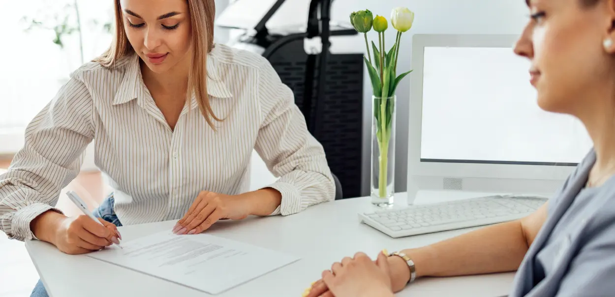 A woman in a striped shirt signing a document at a table, while another woman sits across from her, observing. There are flowers in a vase in the background and a computer monitor to the right.