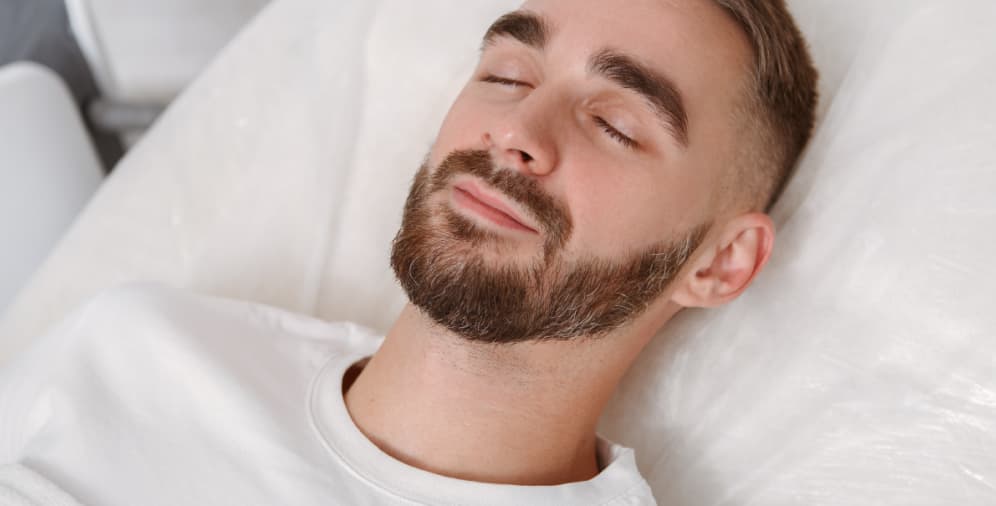 A close-up image of a man with short hair and a beard lying on a bed with his eyes closed, appearing relaxed.