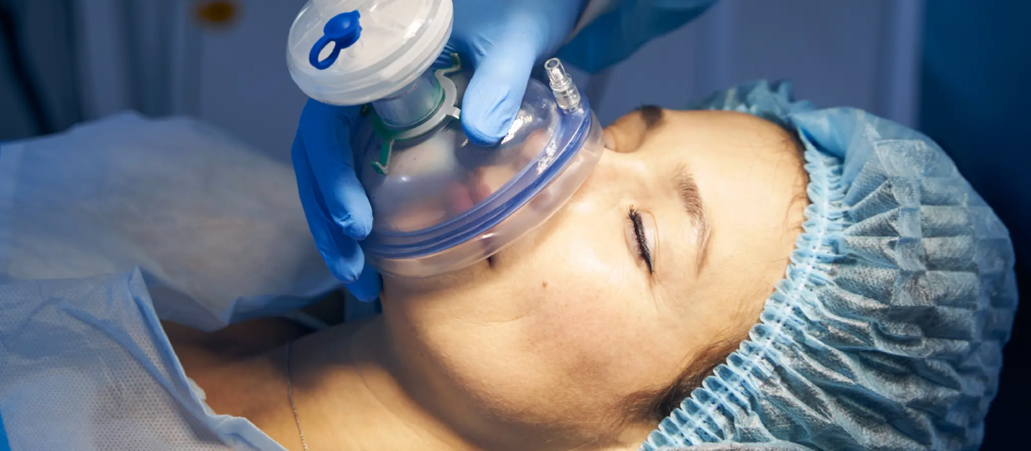 A medical professional in blue gloves holds an anesthesia mask over a patient's face, who is lying down with closed eyes, wearing a surgical cap and a hospital gown.