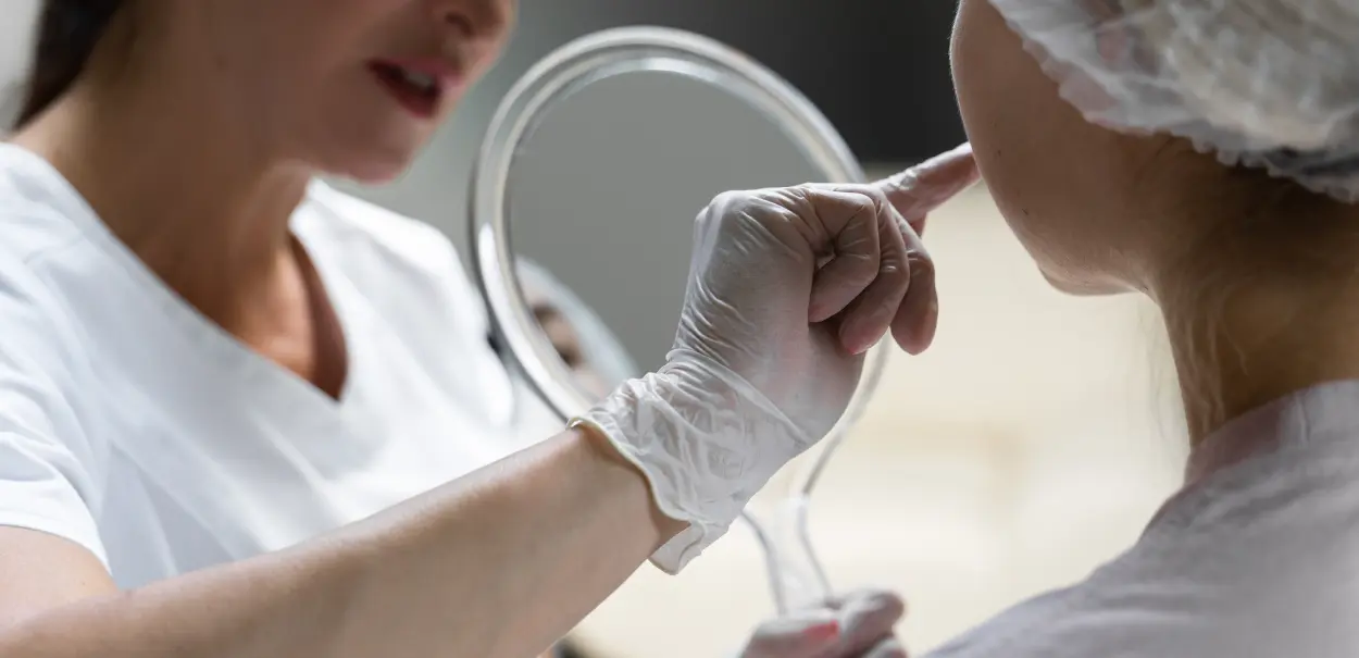 A close-up of a healthcare professional wearing gloves, holding a handheld mirror, pointing toward the face of a patient. The professional is speaking while the patient listens attentively.
